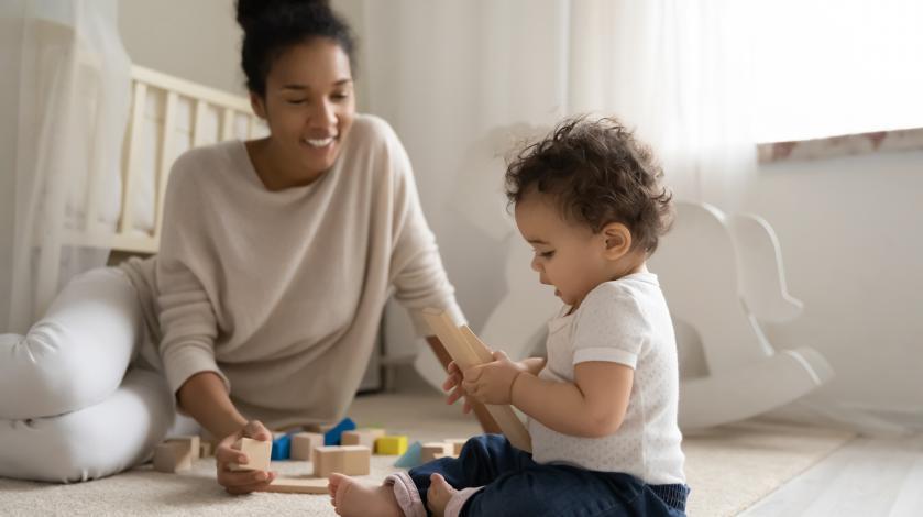 A baby sits on the floor of his nursery next to his mother as he plays with colorful wooden blocks