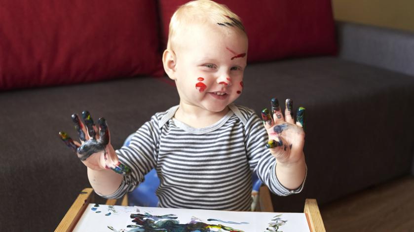 A happy baby covered in paint holds up his hands after finger painting