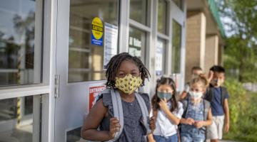 A group of happy-looking children stands outside of their classroom preparing for the first day of school. Wearing masks due to COVID-19.
