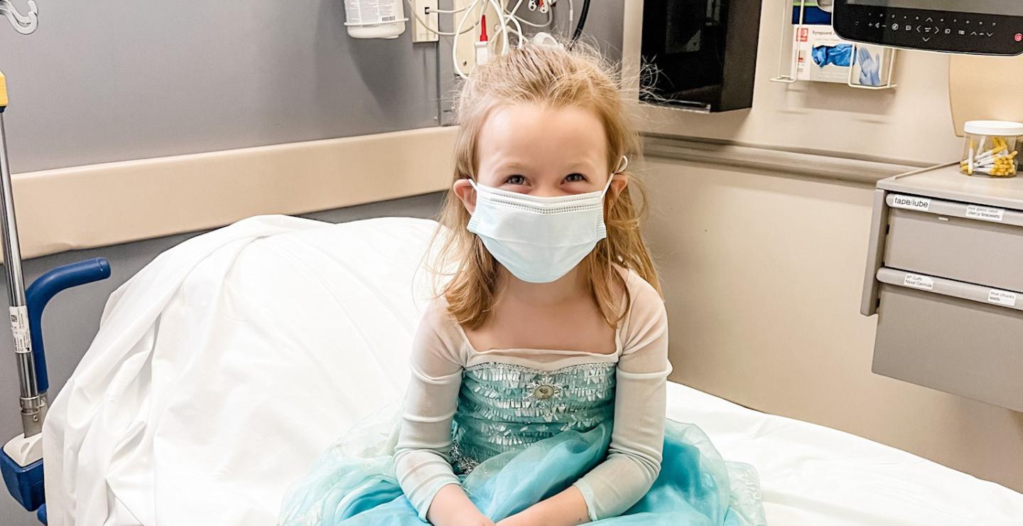 Bizzy, a young patient at South Shore Hospital, smiles as she sits on a bed wearing her Elsa princess gown.