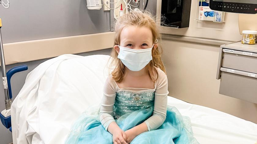 Bizzy, a young patient at South Shore Hospital, smiles as she sits on a bed wearing her Elsa princess gown.