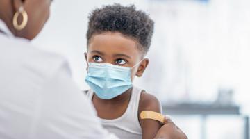 A young boy wearing a mask looks up at his doctor as she puts a band aid on his arm after giving him a vaccine