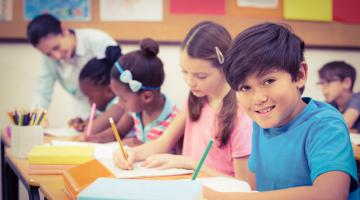 A young boy sitting at his desk in school writing on a piece of paper. Other classmates are doing similarly in the background.