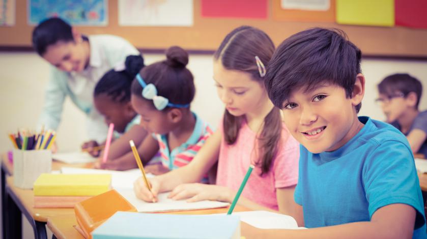 A young boy sitting at his desk in school writing on a piece of paper. Other classmates are doing similarly in the background.