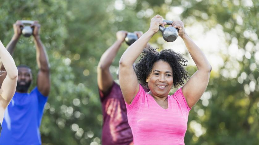 A woman smiles as she holds a weight over her head during an outdoor exercise class.
