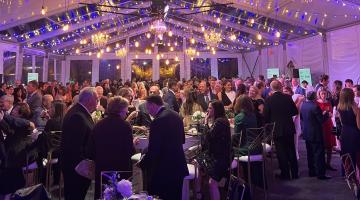 Guests in formal attire mingle beneath string lights in a white tent the Moonlight & Miracles Gala in Weymouth, Massachusetts.