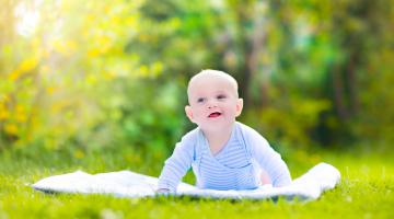 A happy baby boy does tummy time on a blanket outside in a green scene