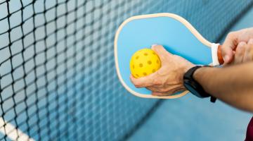 A view of a person holding a yellow pickleball in front of a blue pickleball paddle.