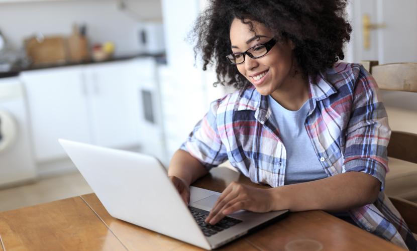 A young woman sits at her dining room table working on a white laptop, smiling while she looking at the screen