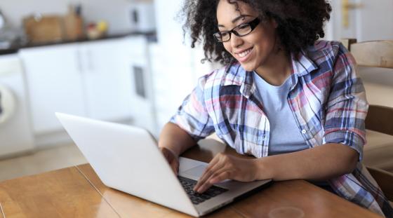 A young woman sits at her dining room table working on a white laptop, smiling while she looking at the screen