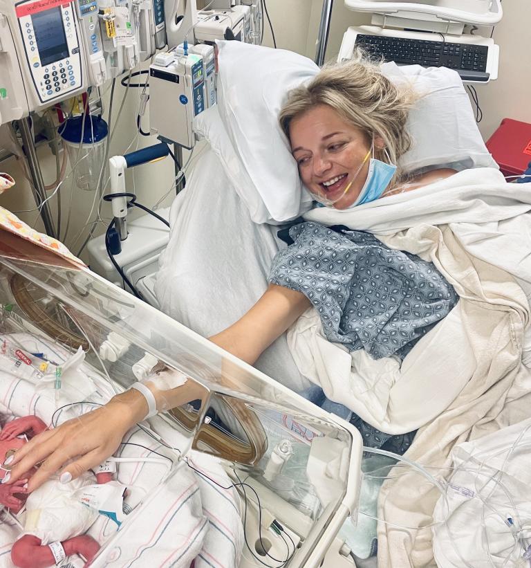 Baby Monroe lies in a NICU bed at South Shore Hospital shortly after birth, as mother Jillian reaches out to touch her, smiling, from her own hospital bed.