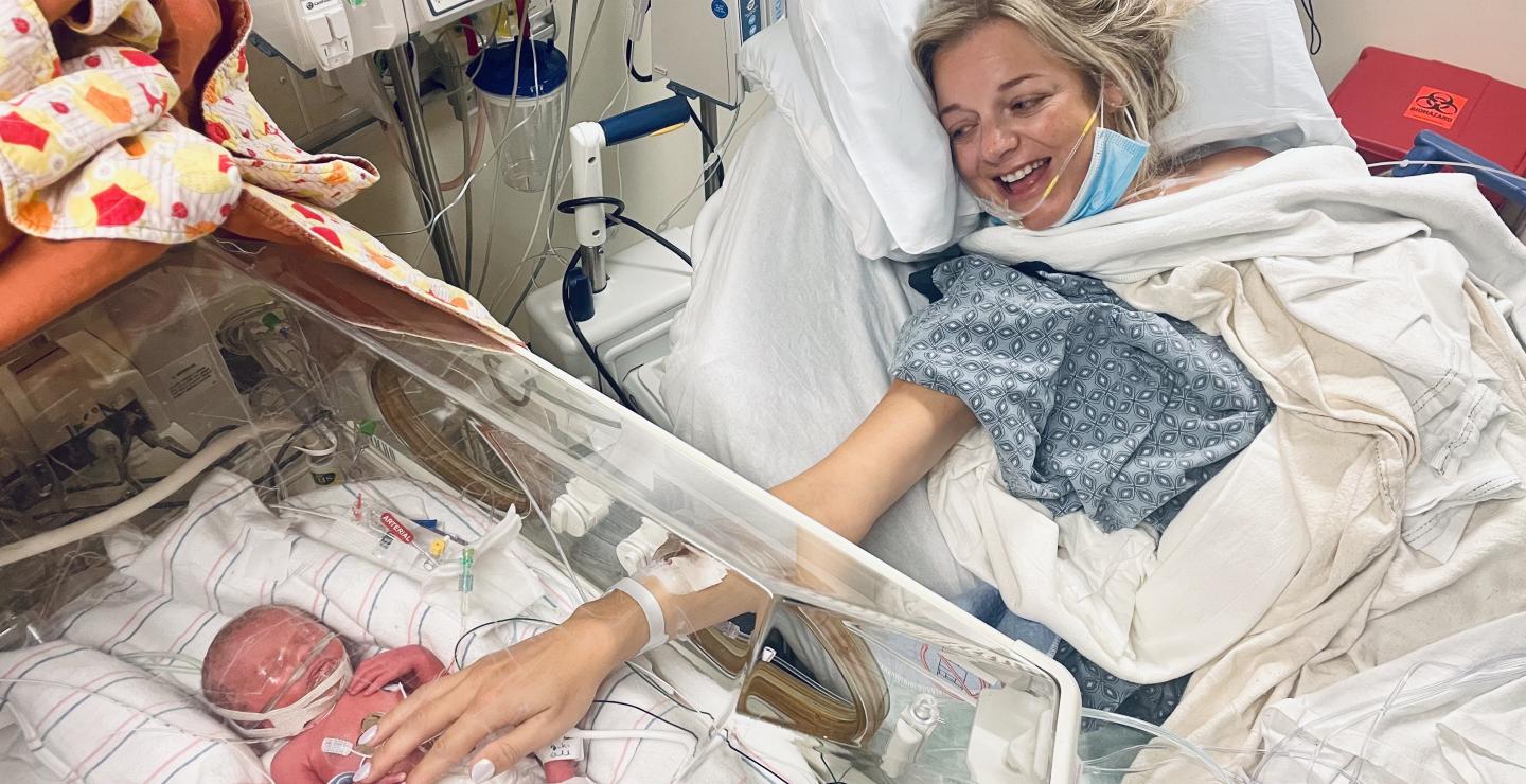 Baby Monroe lies in a NICU bed at South Shore Hospital shortly after birth, as mother Jillian reaches out to touch her, smiling, from her own hospital bed.