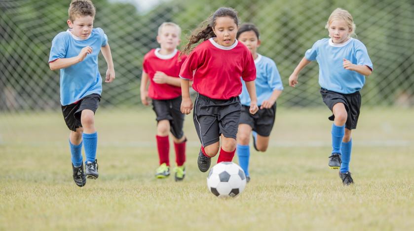 A girl in a red shirt and soccer uniform kicks a ball as four other players chase behind her in a youth soccer game.