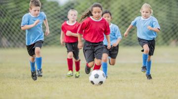 A girl in a red shirt and soccer uniform kicks a ball as four other players chase behind her in a youth soccer game.