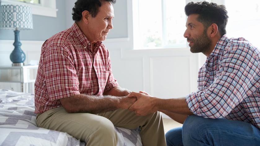 An older man smiles sitting on the edge of his bed as he talks to his adult son, who is holding his hand.