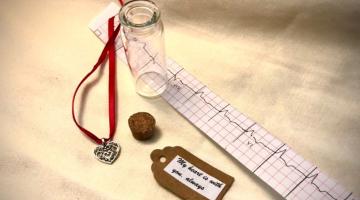 A metal heart pendant on a red ribbon, a cork, an empty class vial, a tag reading "my heart is with you, always," and a printed EKG heartbeat.