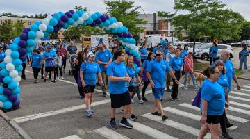 Walkers wearing light blue shirts pass under a white, blue, and purple balloon arch at the start of the 2022 Walk for Hospice