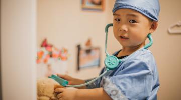A young Asian boy dressed as a doctor plays pretend by putting his stethoscope on a stuffed animal